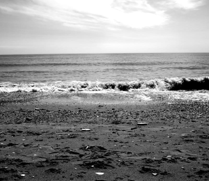 Grayscale photo of the sea with the horizon in the back and sandy beach in the front. Some small waves are forming where the sea meets the beach.